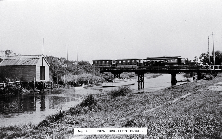 An electric tram crosses the New Brighton bridge with a barge moored underneath 